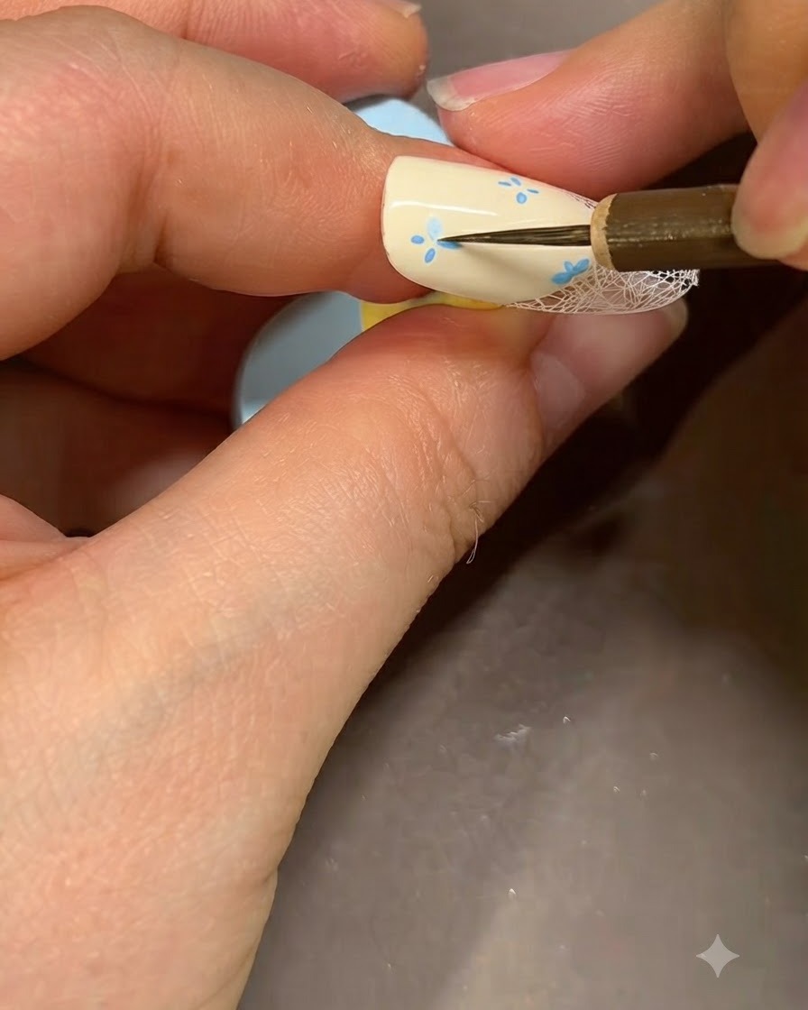 A fine nail art brush adding detailed blue accents to the center of a painted flower on a beige nail.
