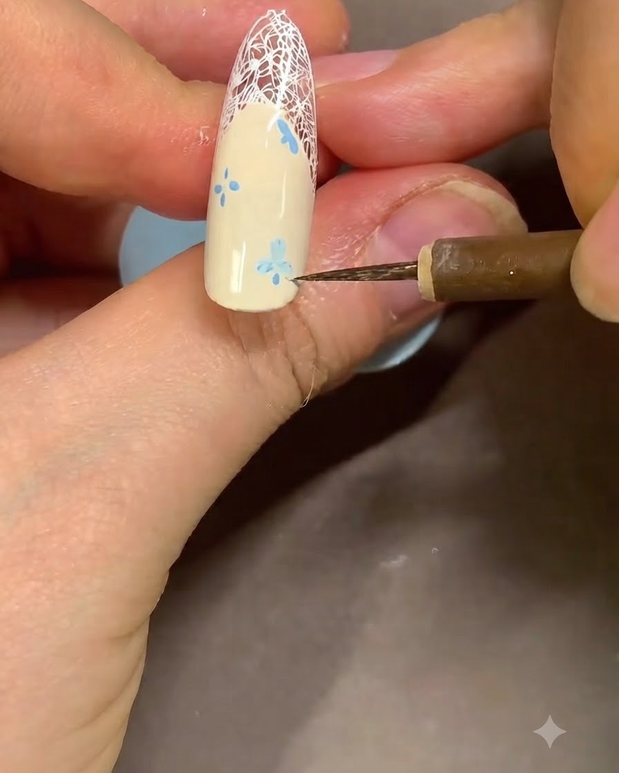 A close-up of a nail artist using a fine detail brush to clean up the edges of a blue flower design near the French tip.