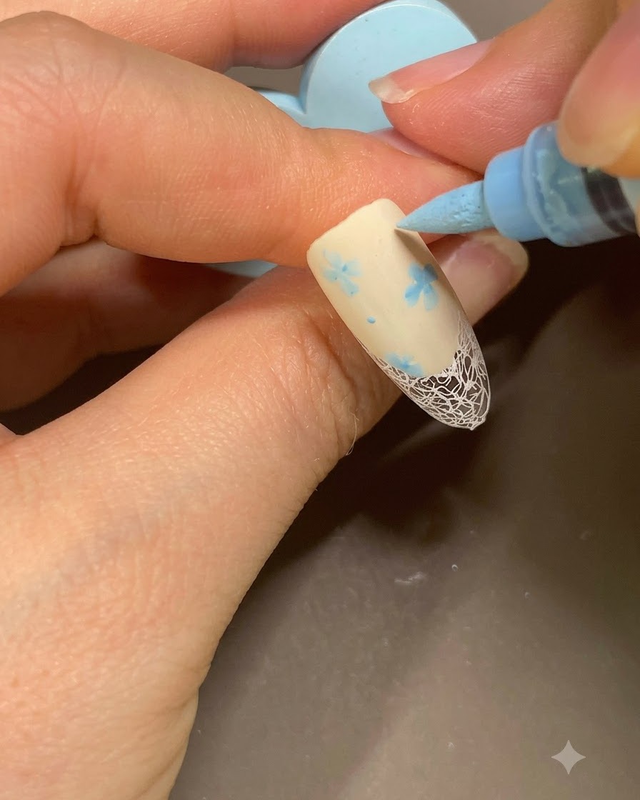 A blue nail art marker adding final small dots next to a floral pattern on a beige French manicure.