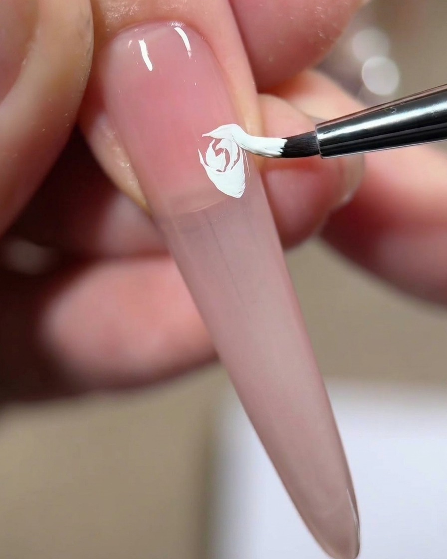 A nail brush pressing and dragging white gel to add a fourth layered petal to a painted white rose on a pink nail.