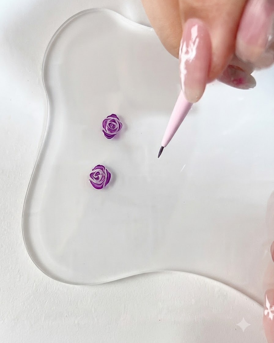 A close-up view of a fine liner brush drawing a small curved purple petal hook on a clear palette.