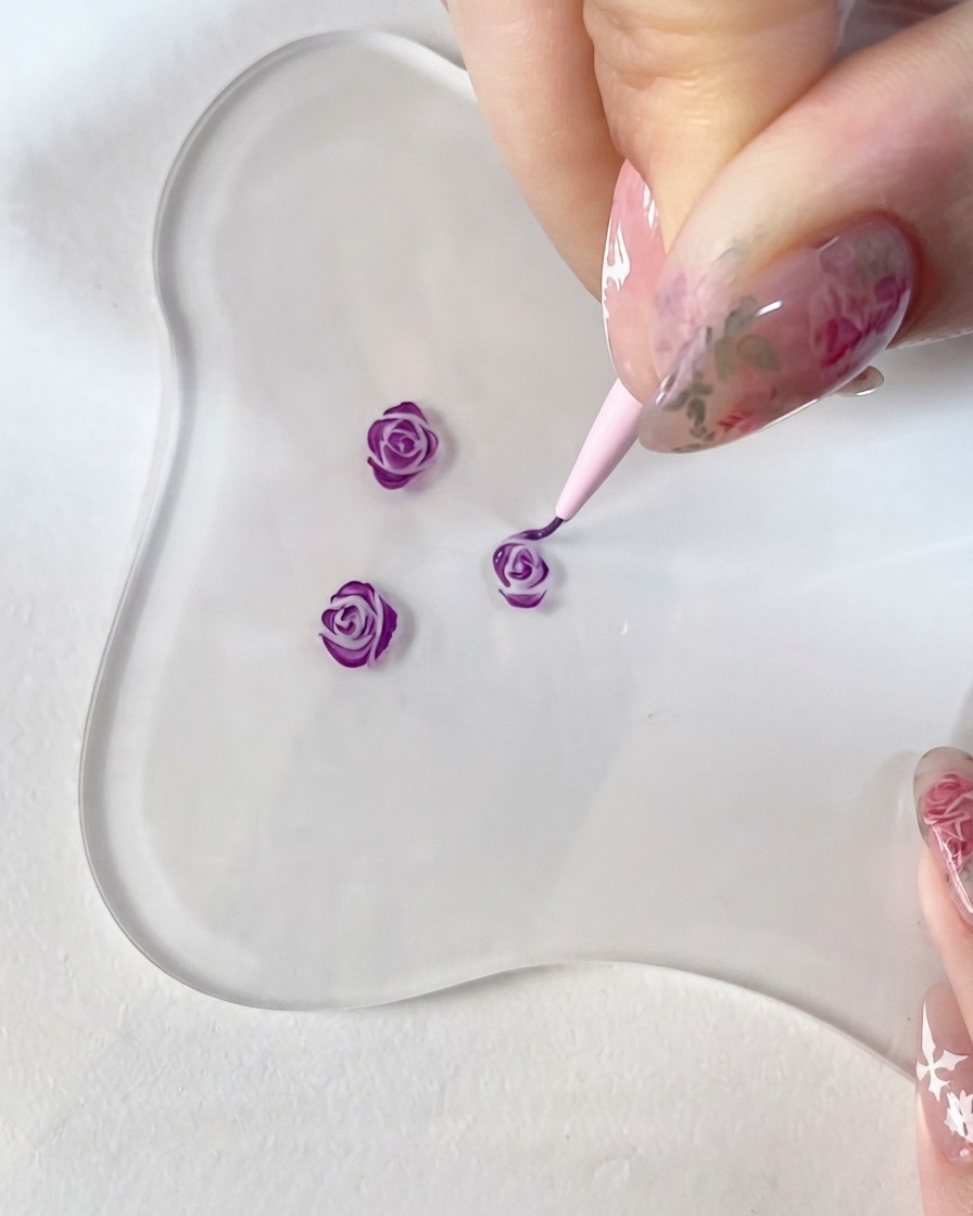A close-up of a hand holding a fine detail brush, meticulously painting dark purple accents onto a small 3D rose design on a clear glass palette.