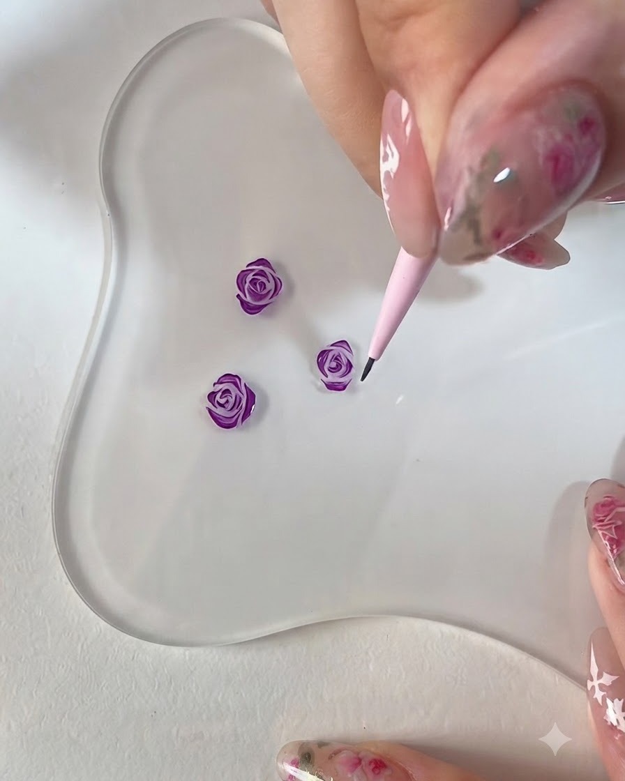 A close-up of a hand using a pink detail brush to refine the edges of a 3D painted purple rose on a clear palette.
