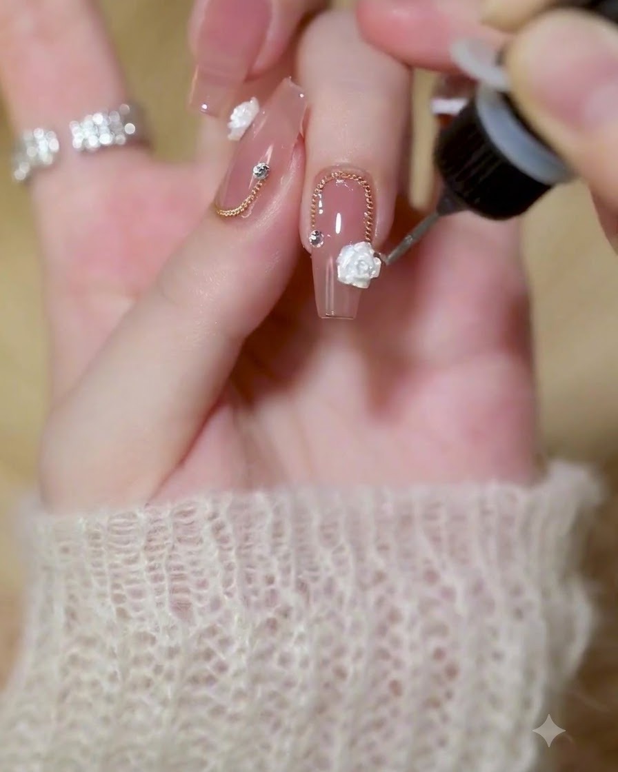 A fine detail brush applying clear builder gel around the edges of a 3D white rose and gold chain on a polished nail.