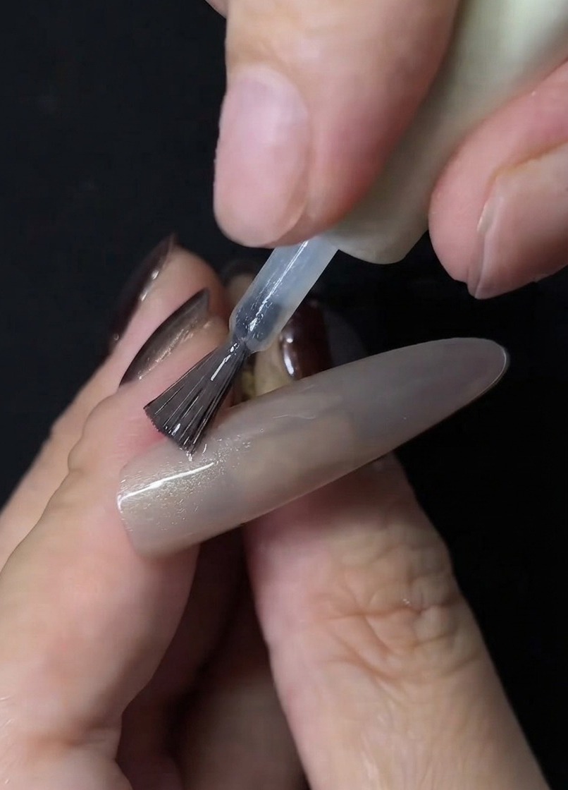 A nail brush applying clear cat-eye gel polish onto the surface of a nude nail extension.