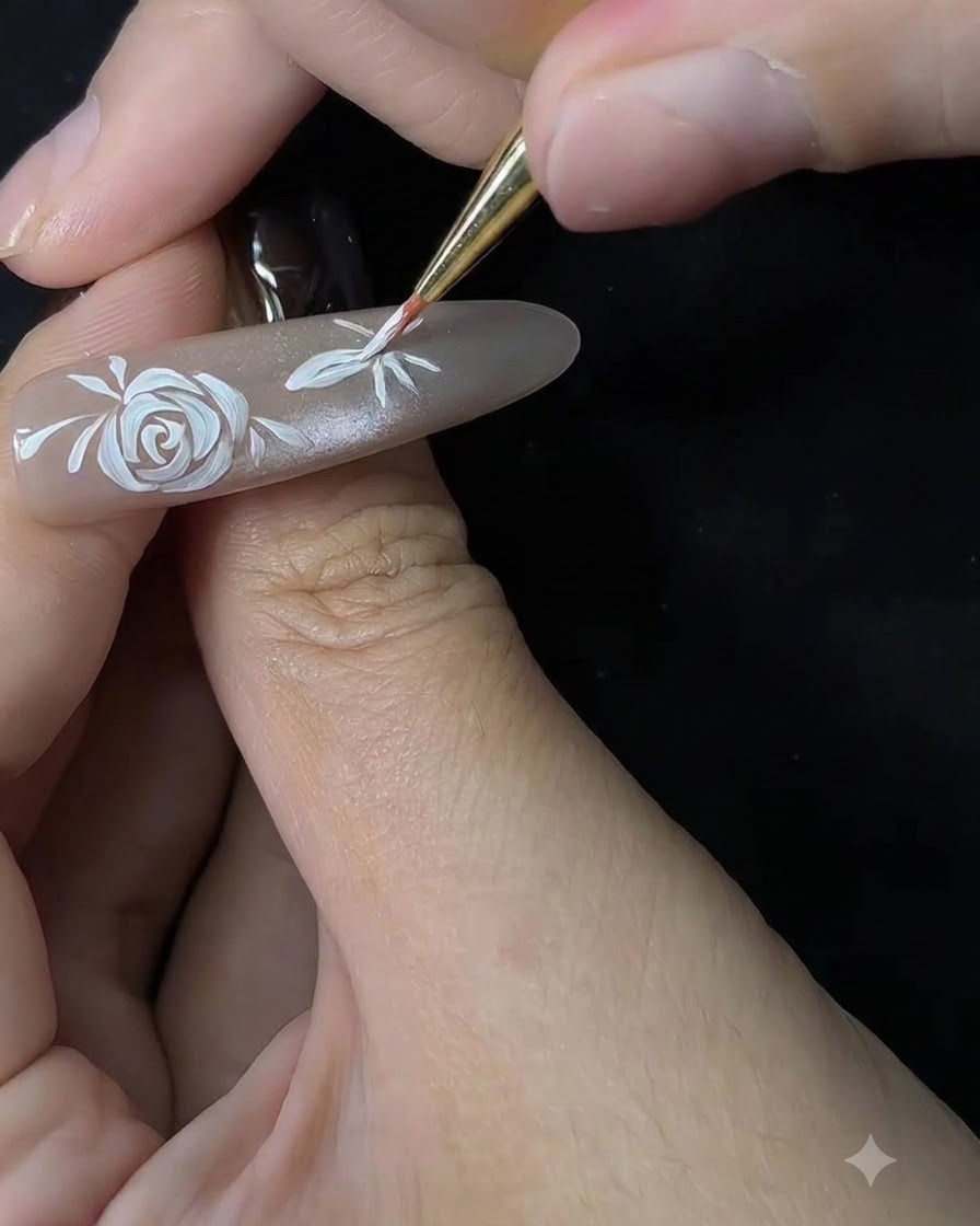 A close-up shot of a fine brush starting to shape the petals of a secondary flower on a nude-colored nail extension.