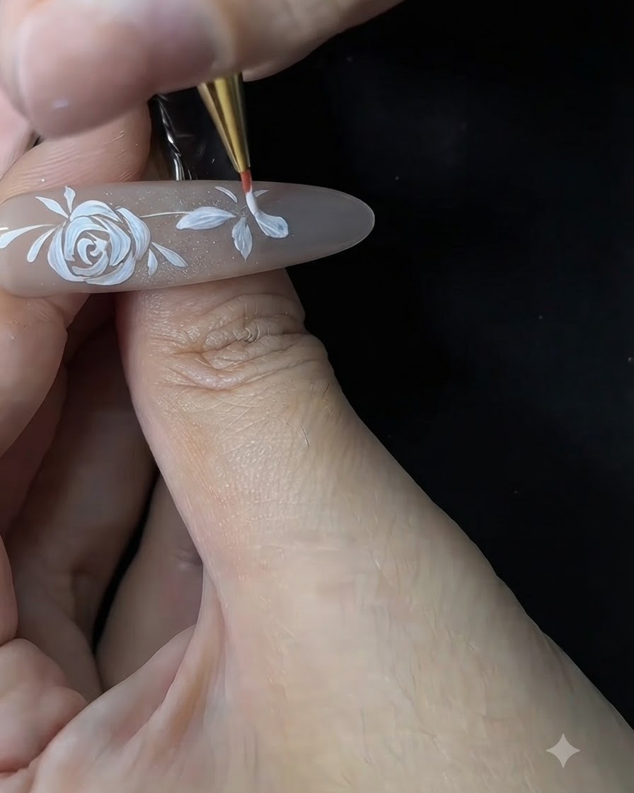 A nail technician using a fine brush to refine and thicken the white petals of a small flower on a gel nail extension.