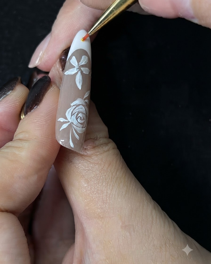 A close-up of a hand using a fine detail brush to paint small white accent vines around a white hand-painted rose on a nude gel nail.