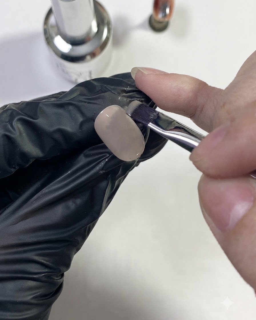 A gloved hand brushing a nude base color onto a nail tip.