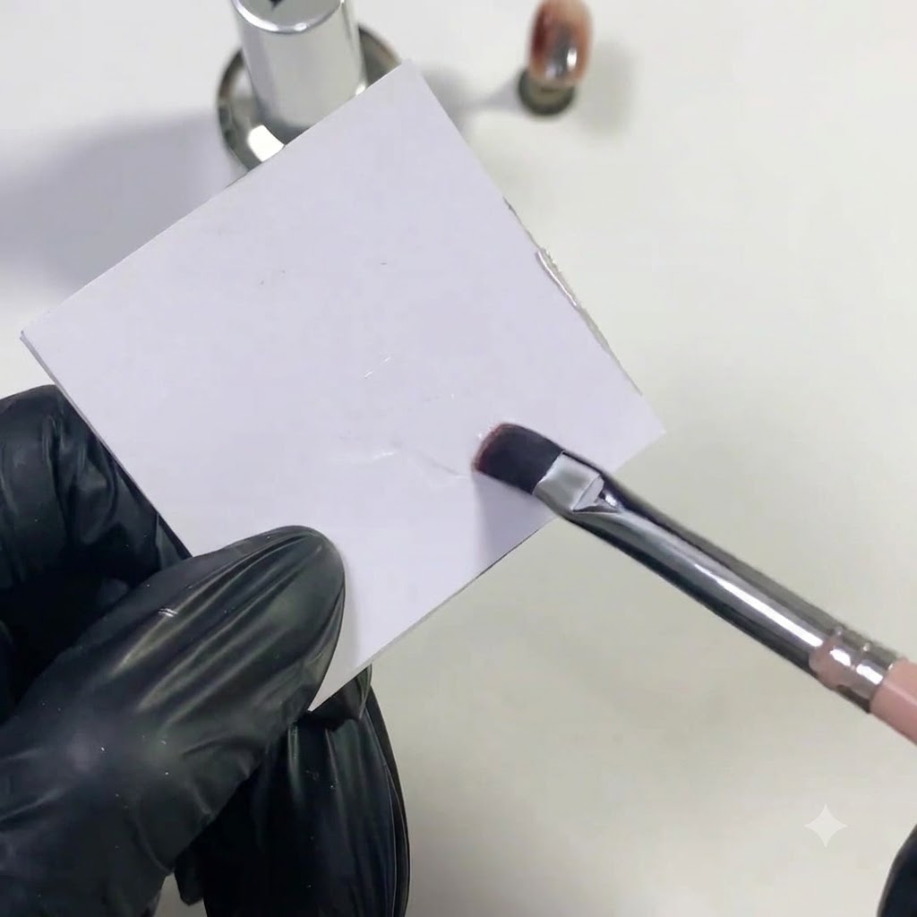 A flat nail brush dabbing clear base gel on a white palette surface.