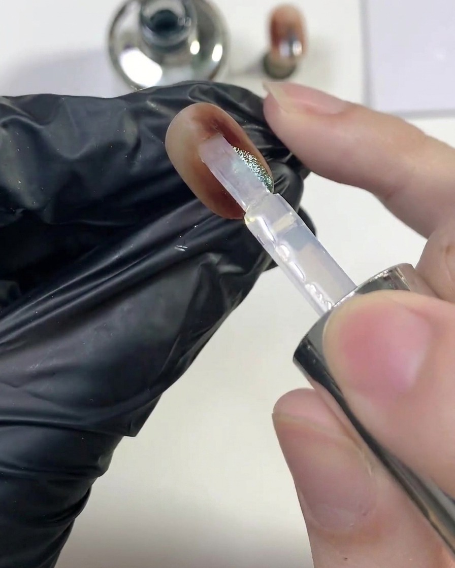 A brush gliding a generous layer of glossy clear top coat over the finished amber cat-eye nail.