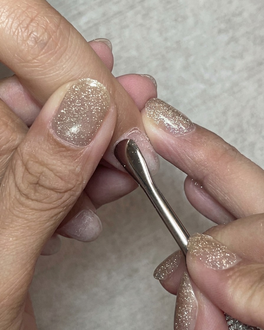 A close-up of a steel cuticle pusher gently pushing back the cuticles on a natural fingernail.