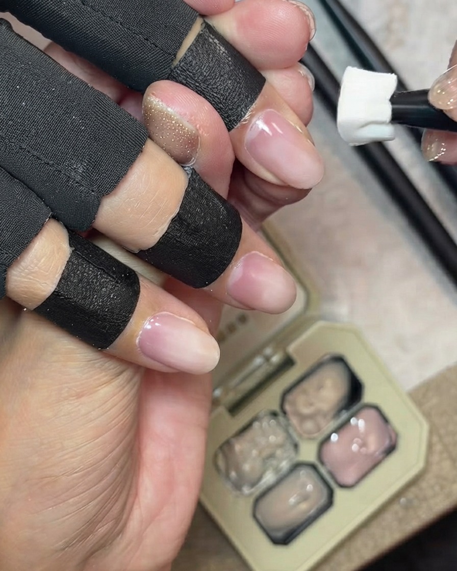A sponge tool dabbing milky nude cover gel across the natural free edge of a fingernail, with a gel palette in the background.