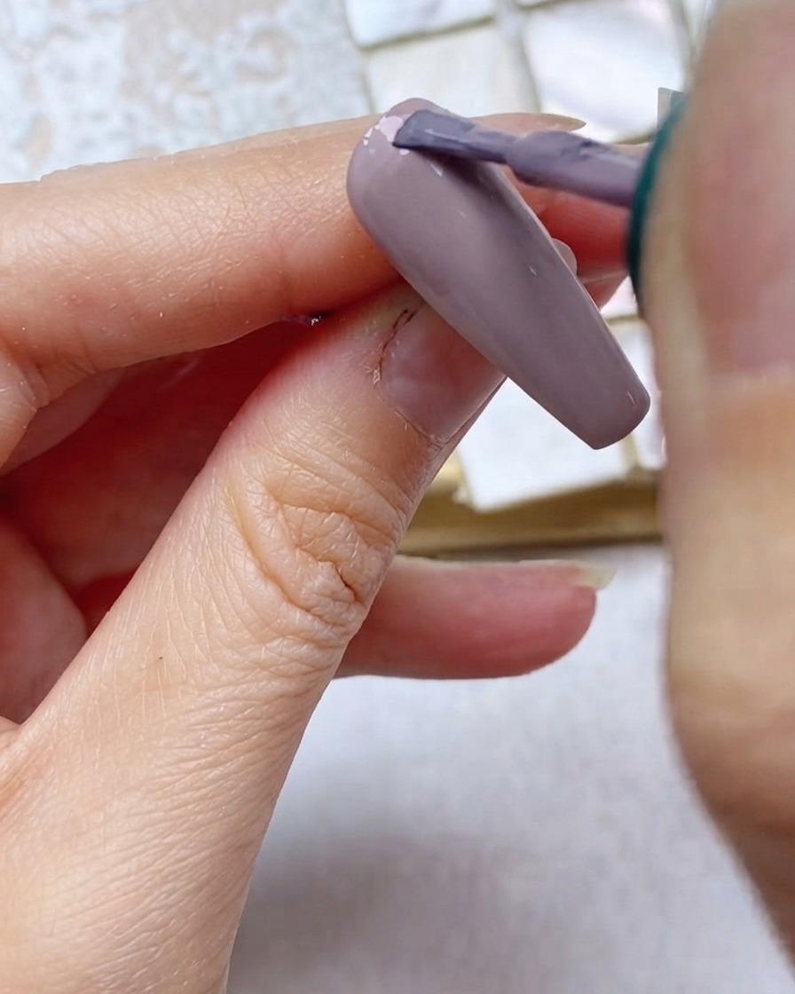 A brush applying grey-purple gel polish to the top one-third of the nail near the cuticle.