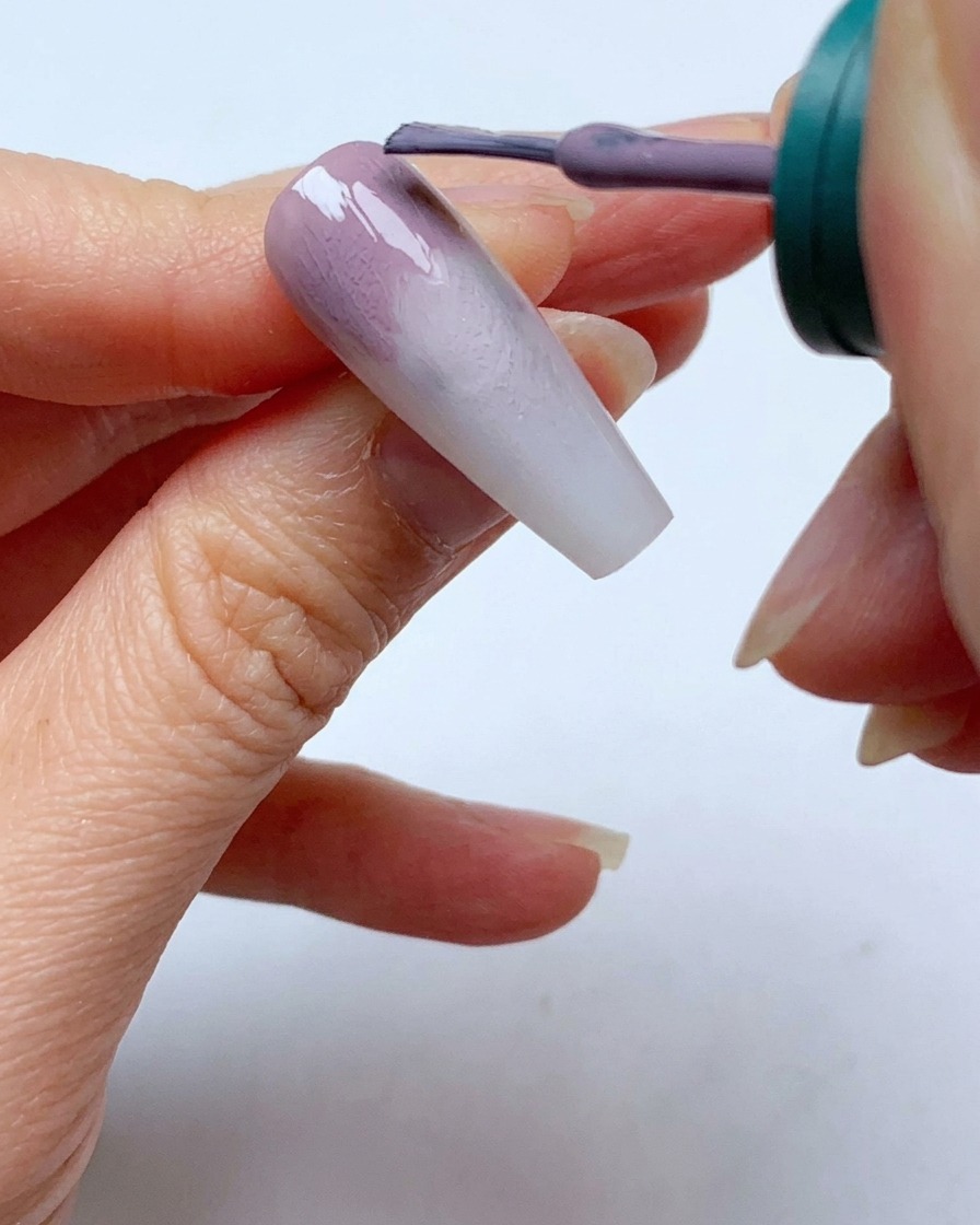 A nail polish brush applying a second layer of grey-purple gel near the cuticle of a nail.