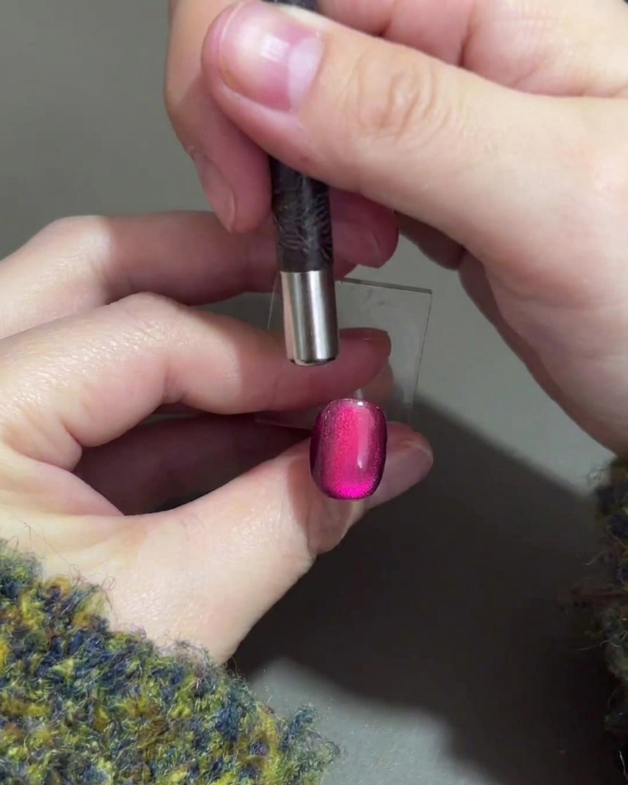 A hand holding a cylindrical magnet over the top tip of a purple gel nail to push the magnetic highlight downwards.