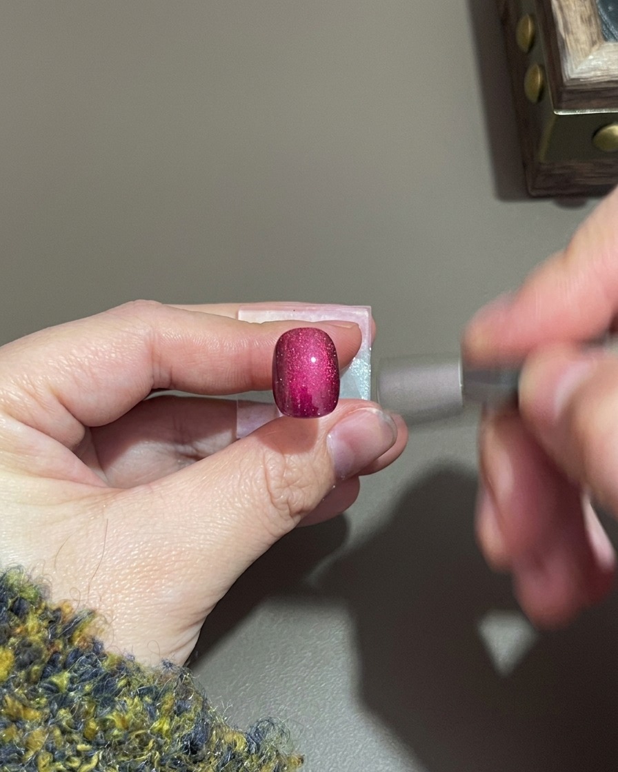 A hand holding a small cylindrical magnet near a purple cat-eye gel nail.