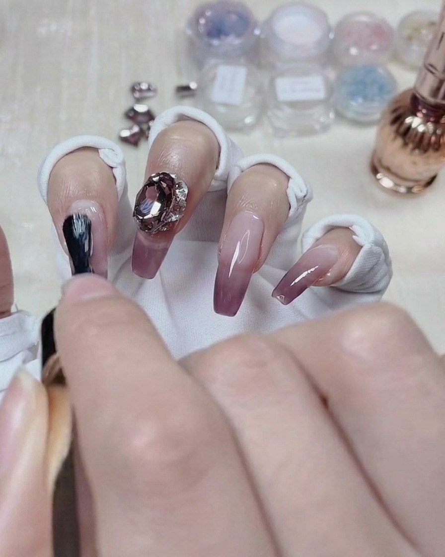 A nail artist applying top coat around the base of a crystal gem on a finished gradient nail.