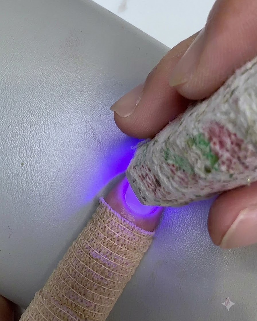 A patterned mini UV/LED lamp held over the tip of a fingernail, curing a blended layer of clear gel.