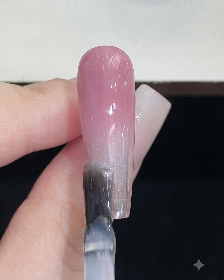 A nail brush applying a clear top coat over a completed pink gradient nail.