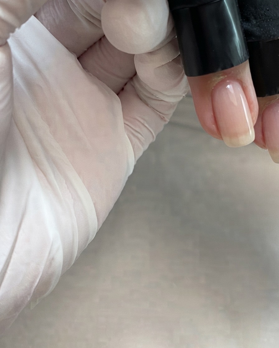 A bottle brush applying a sheer nude pink gel down the center of a fingernail.