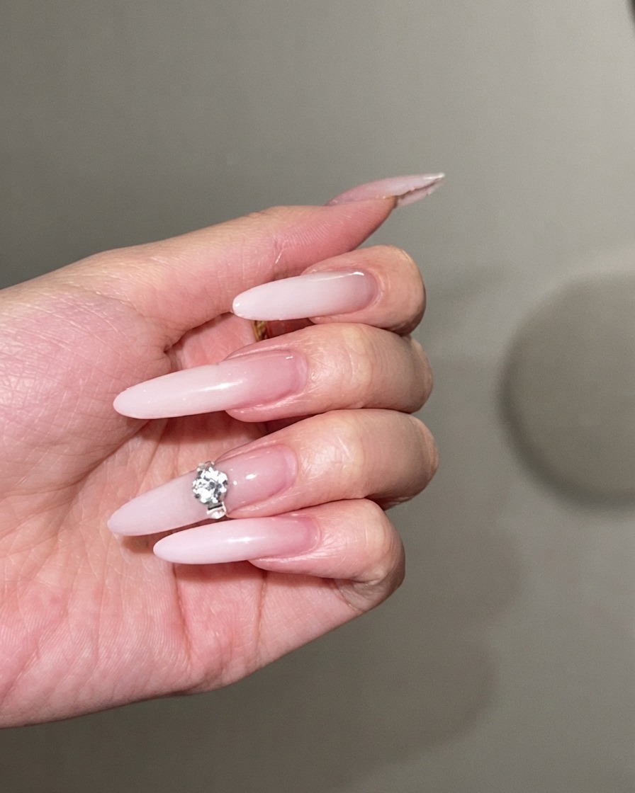 A large sparkling rhinestone being pressed onto the center of a long milky white ombre stiletto nail.