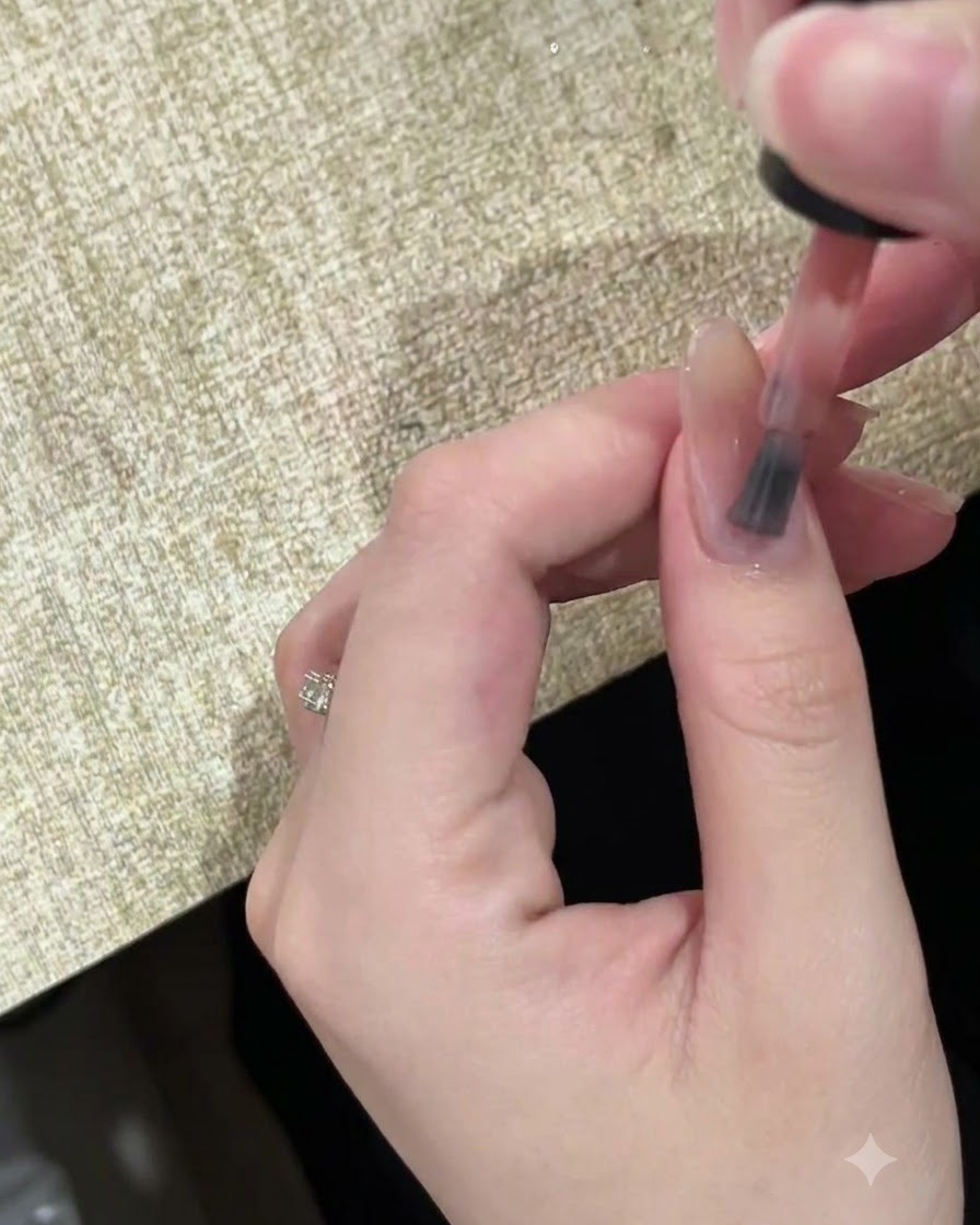 A close-up of a brush applying a sheer nude gel polish to a natural fingernail.