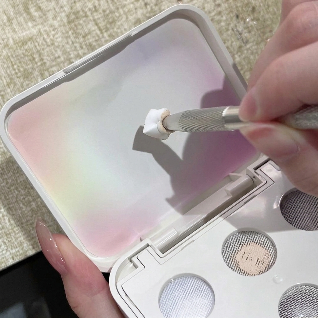 A metal nail tool pressing white solid gel onto the textured grid of a palette lid.