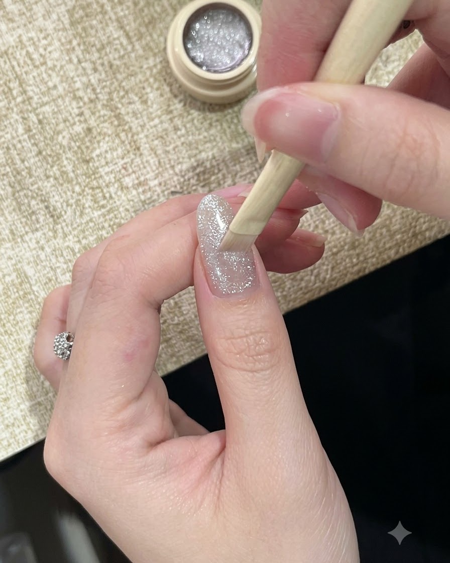 A brush applying a highly reflective, sparkly silver cat-eye gel polish to a fingernail.