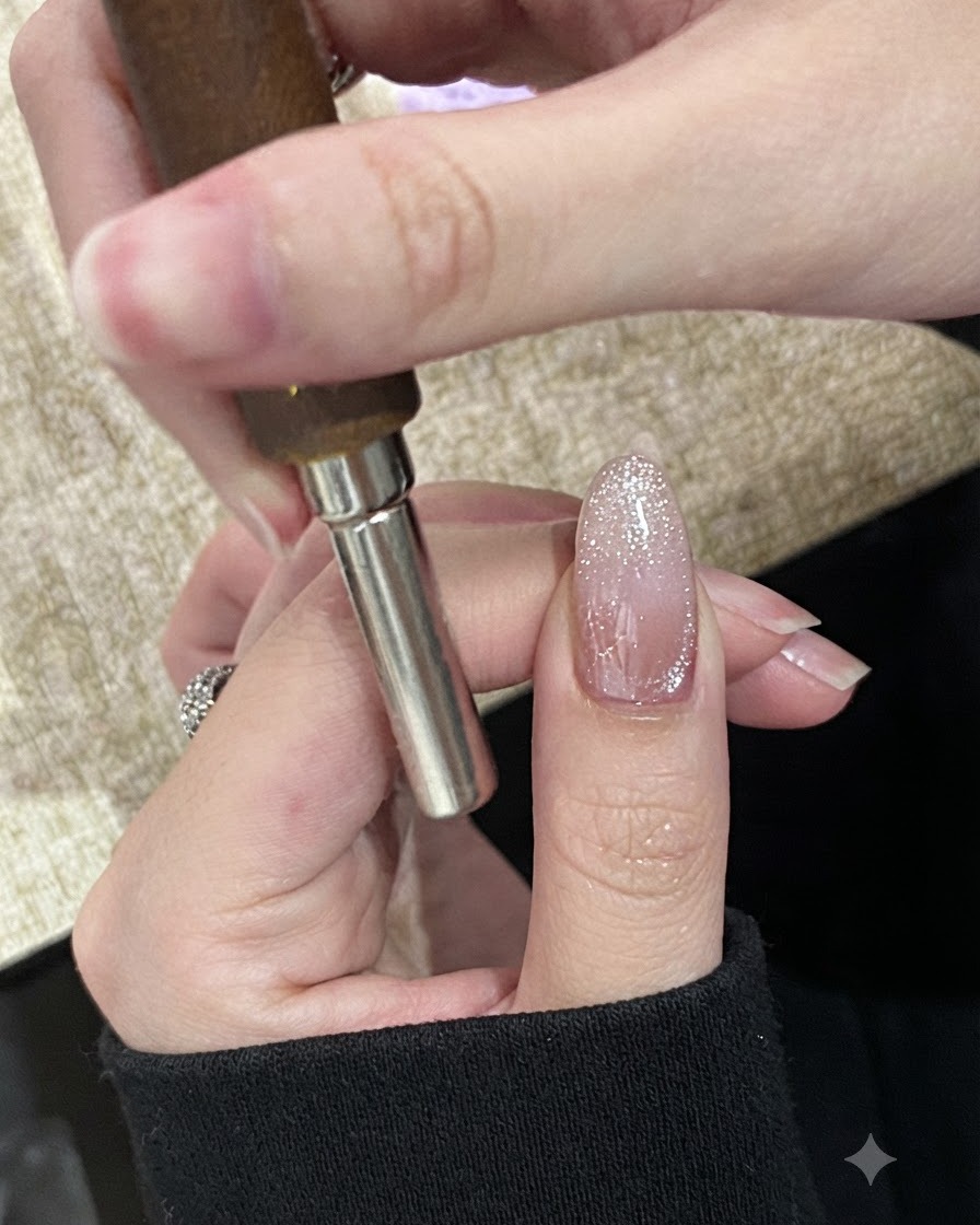 A hand holding a cylindrical magnet horizontally across the tip of a sparkly, un-cured cat-eye gel nail.