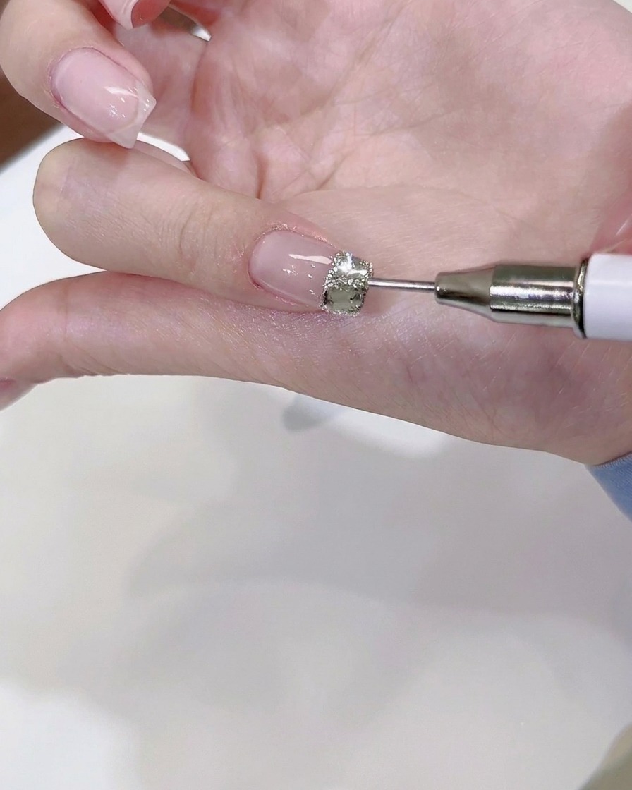 A fine dotting tool placing tiny steel beads and mini crystals around a large central rhinestone on a fingernail.