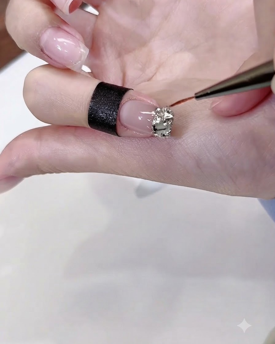 A thin nail art brush applying clear diamond gel glue precisely around the edges of a cluster of rhinestones on a polished fingernail.