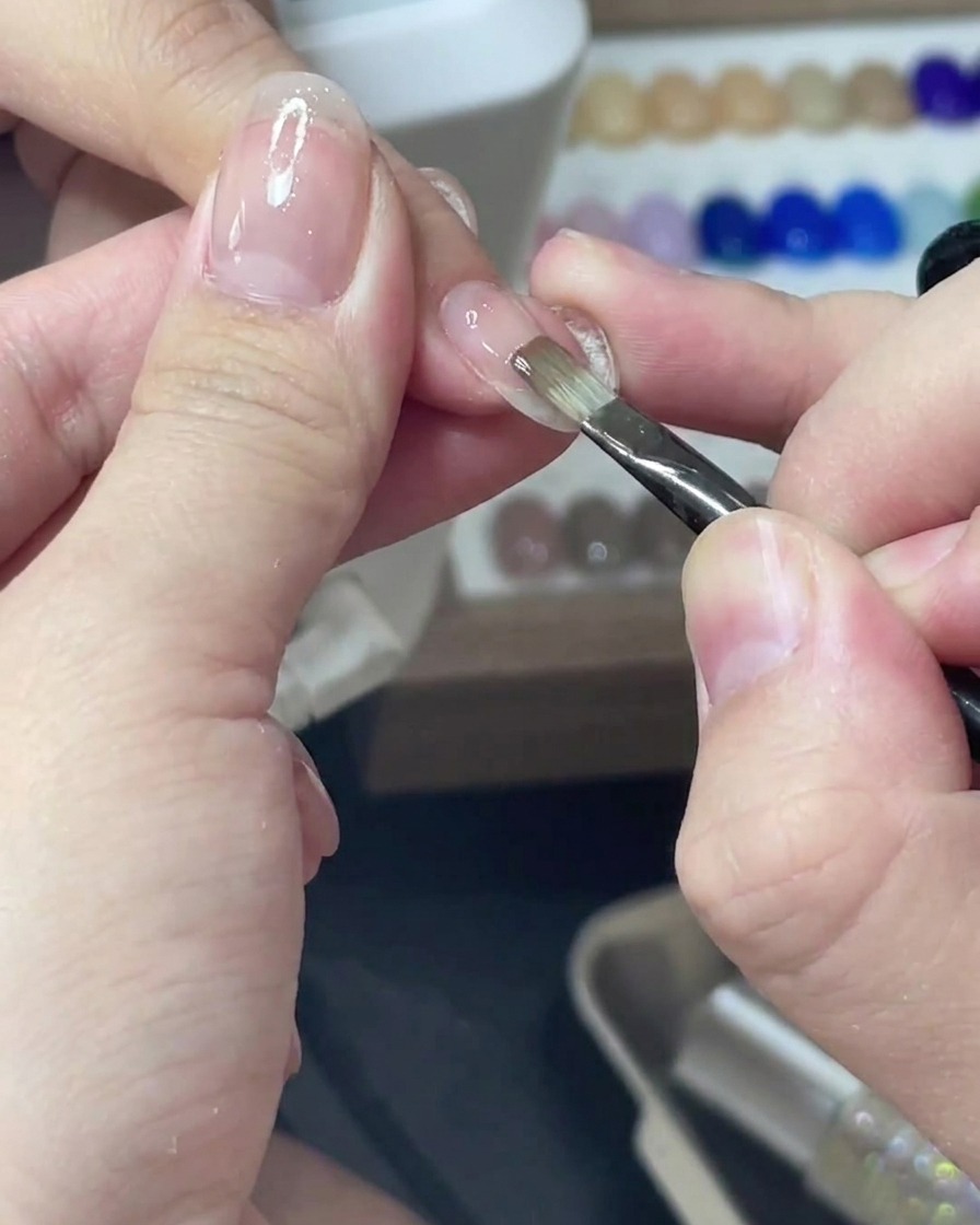 A thin liner brush applying a sheer pink gel polish evenly over a fingernail.