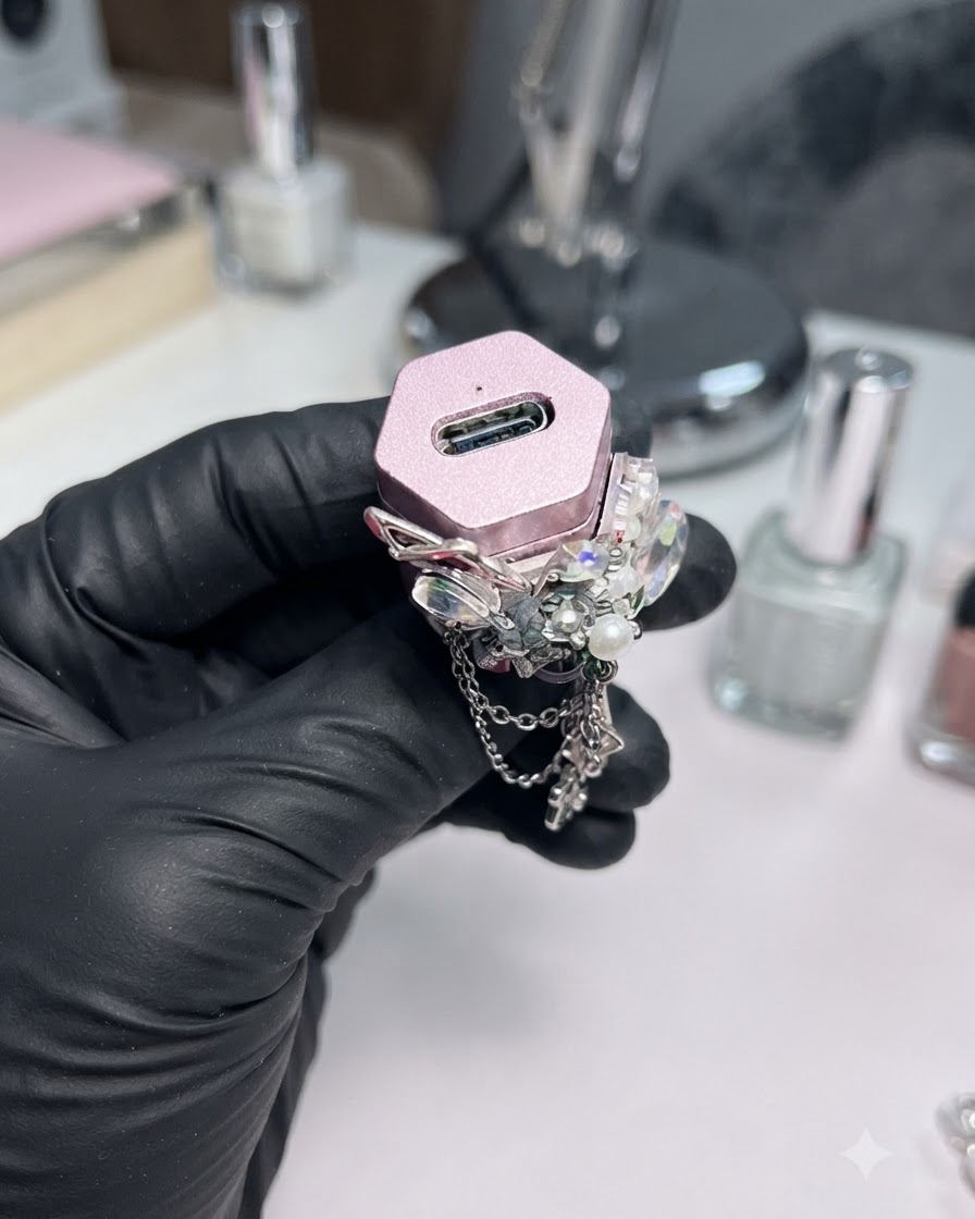 A gloved hand holding a small pink UV/LED lamp decorated with rhinestones.