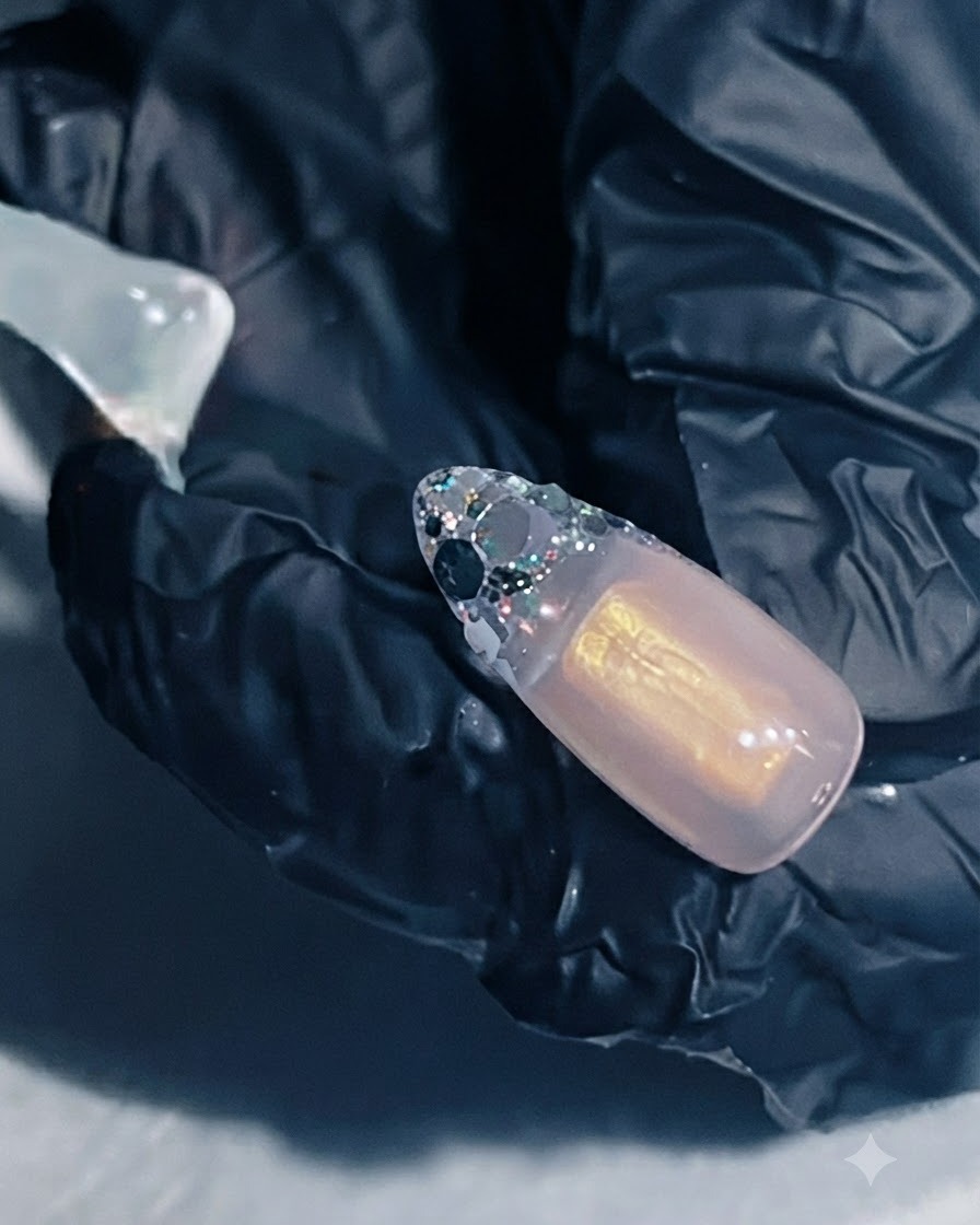 A hand holding a nail extension with reinforcing gel applied over the glitter French area.