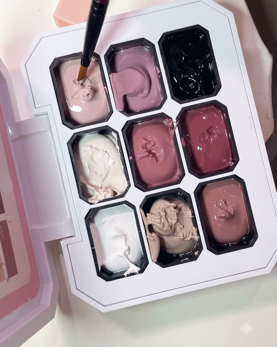 A fine brush picking up nude pink gel polish from a multi-compartment palette.