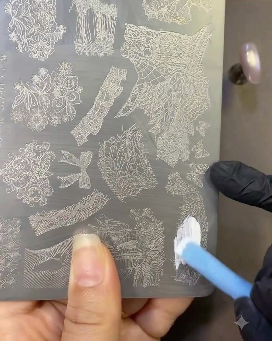 A brush applying white stamping polish onto a metal plate engraved with lace designs.
