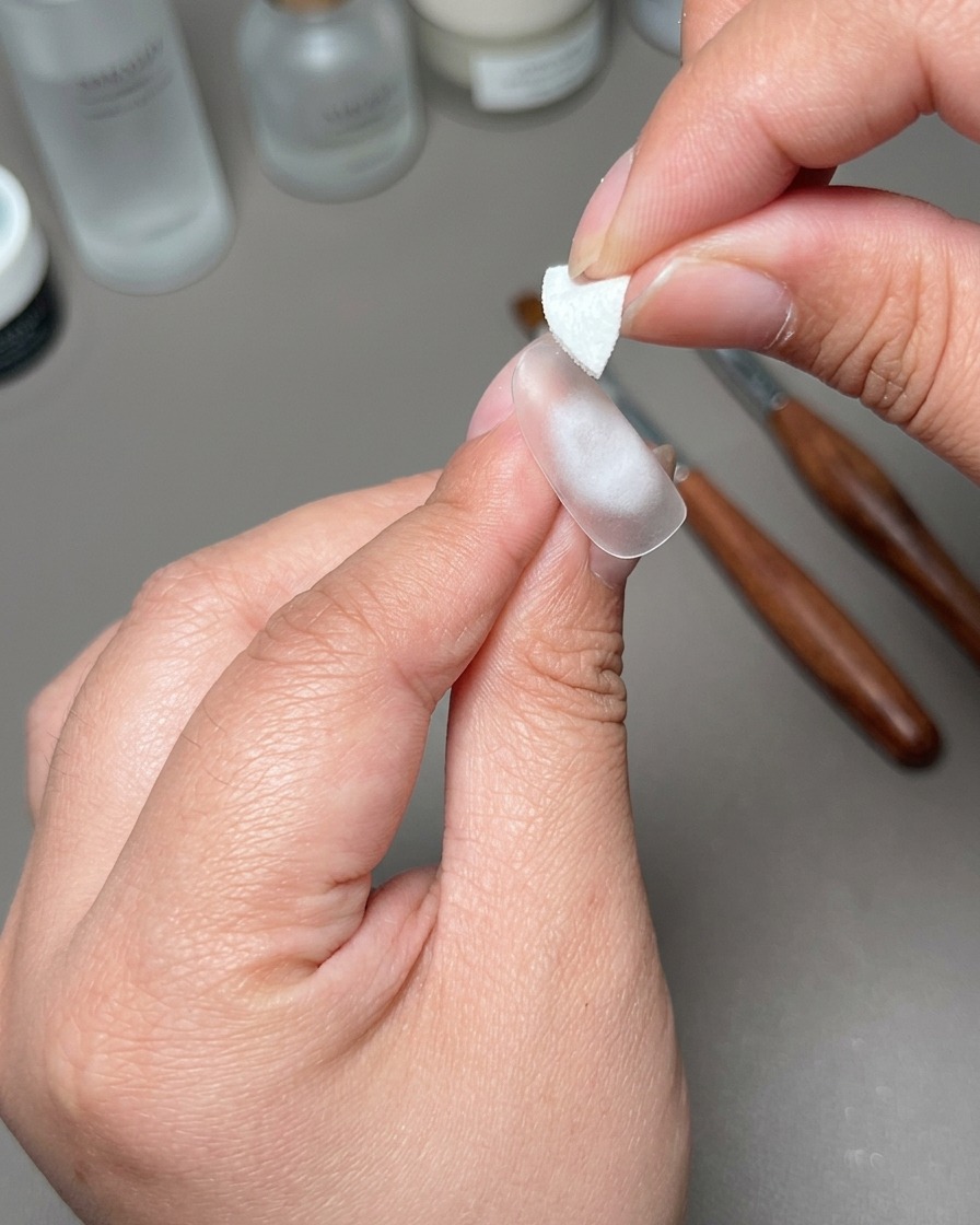 A hand holding a small white adhesive patch over the center of a prepped nail tip.