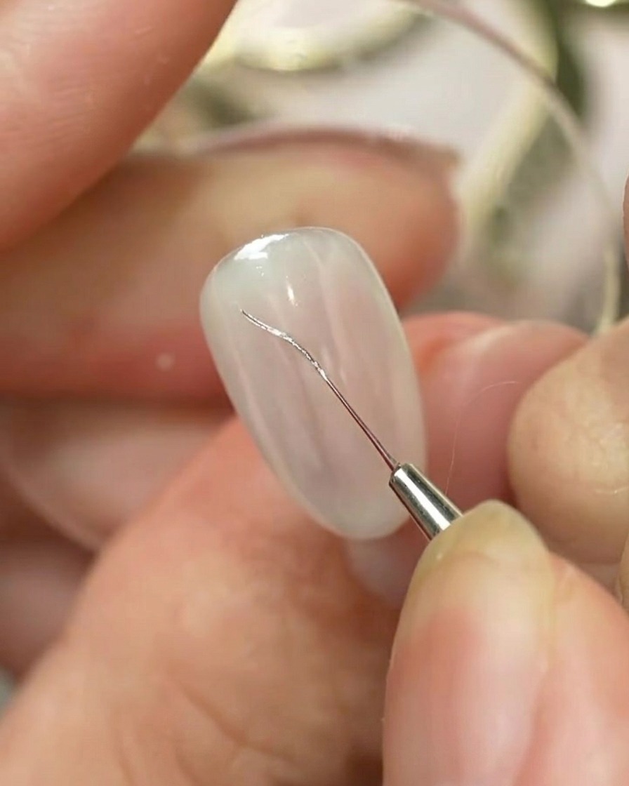 A fine nail art liner brush drawing a silver outline of a bow on a light-colored manicured nail.