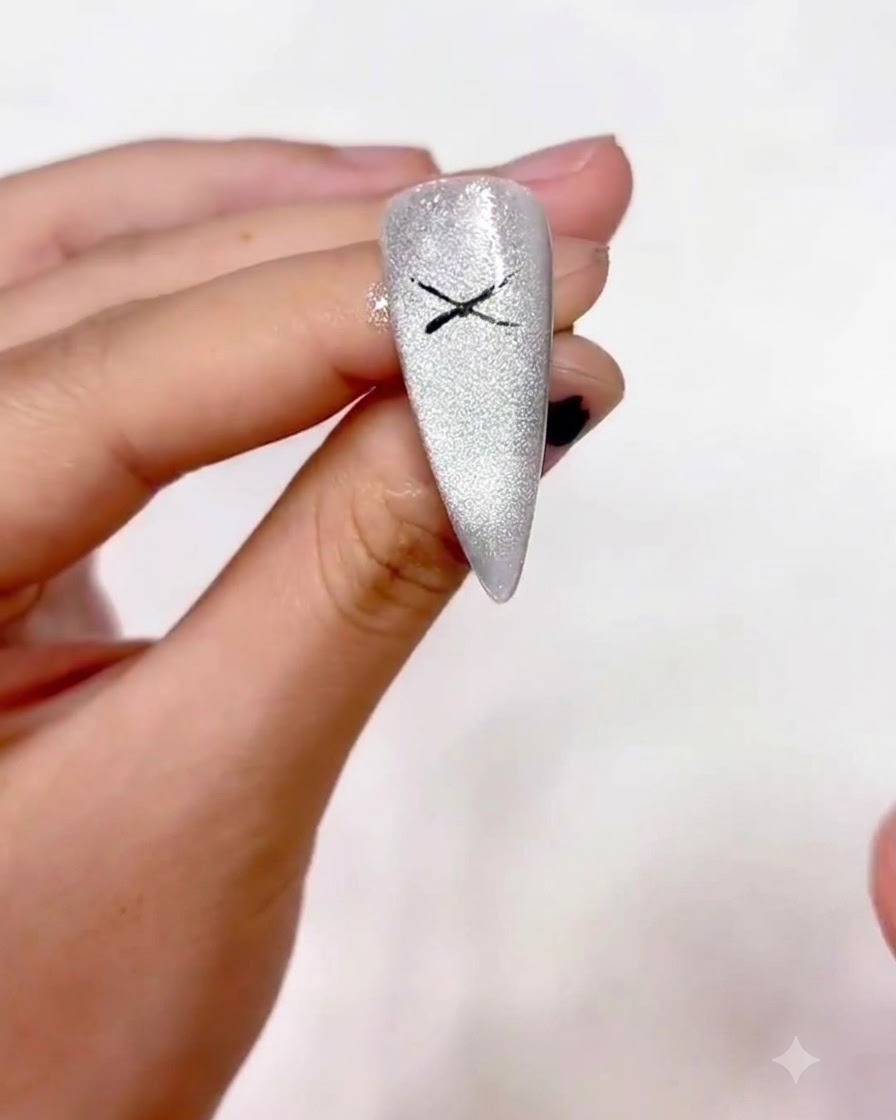 A close-up view of a hand holding a fine liner brush, drawing a black X shape at the center of a shimmery silver cat-eye nail.
