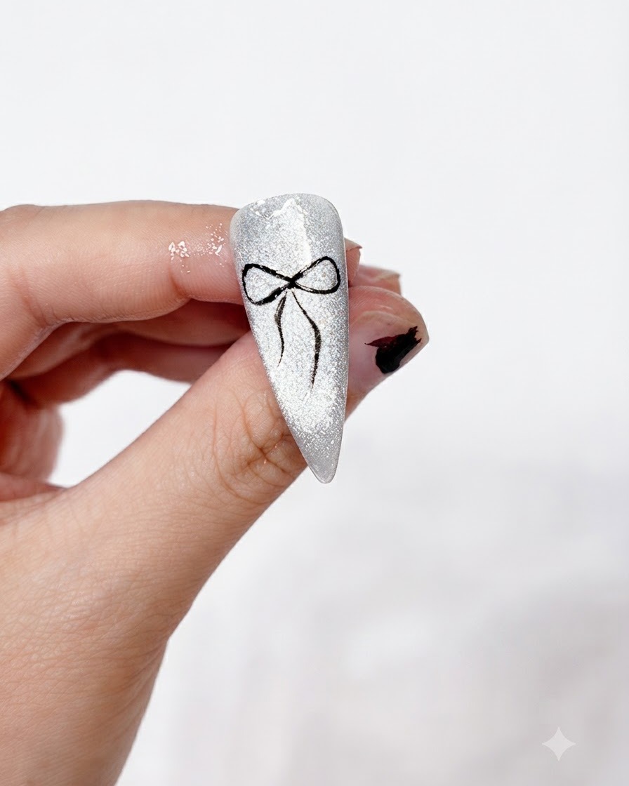 A completed black bow nail art design cleanly painted and set on a sparkling silver cat-eye background, held between fingers.