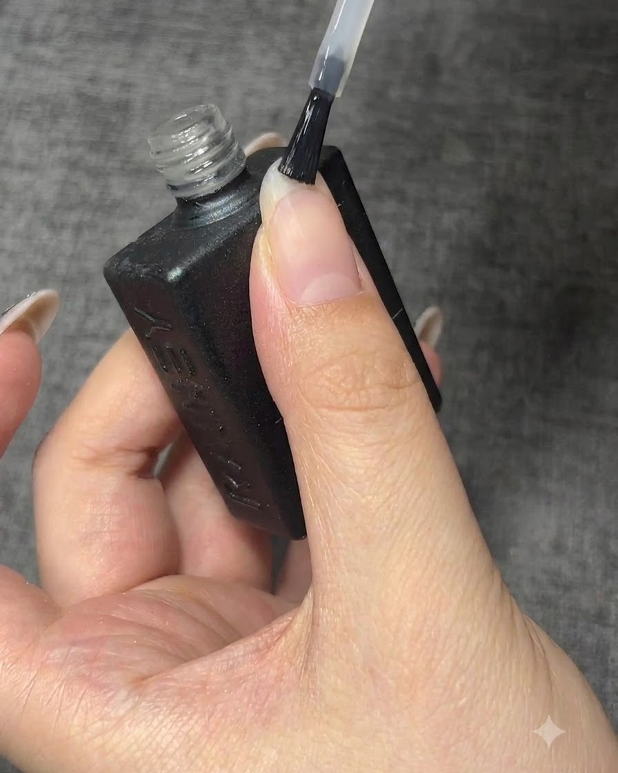 A bottle brush applying clear base coat onto a clean natural fingernail.