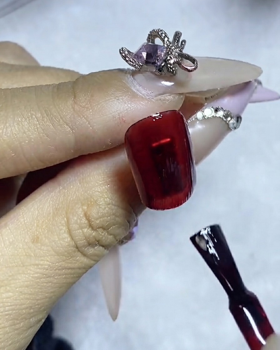 A liner brush applying a precise line of gem glue onto a dark red polished nail surface.