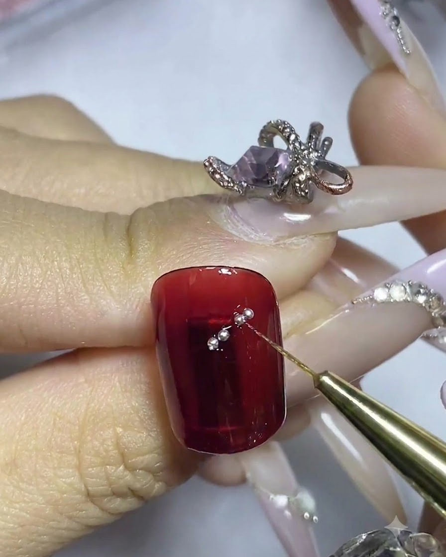 A fine brush placing individual small silver steel beads along the adhesive outline on a red nail.