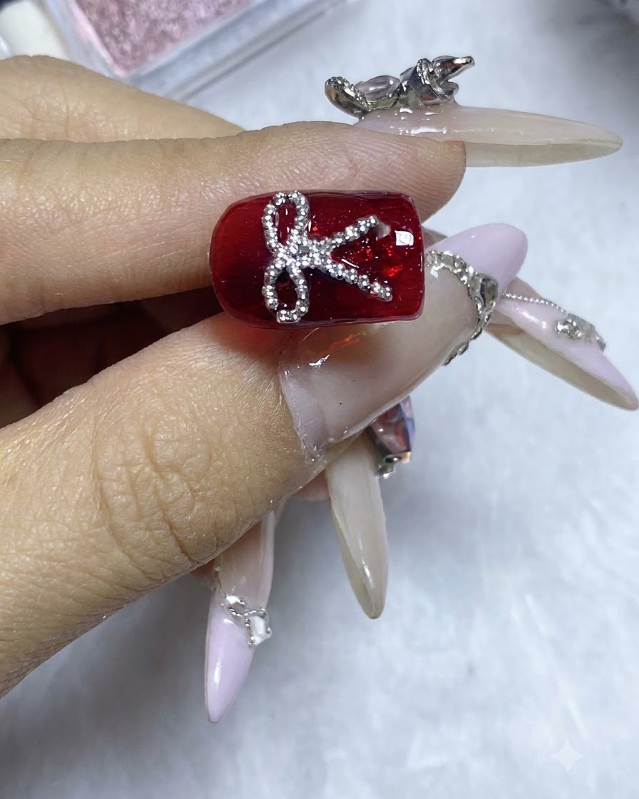 A completed red nail design featuring a sparkling steel bead bowknot under bright studio lighting.