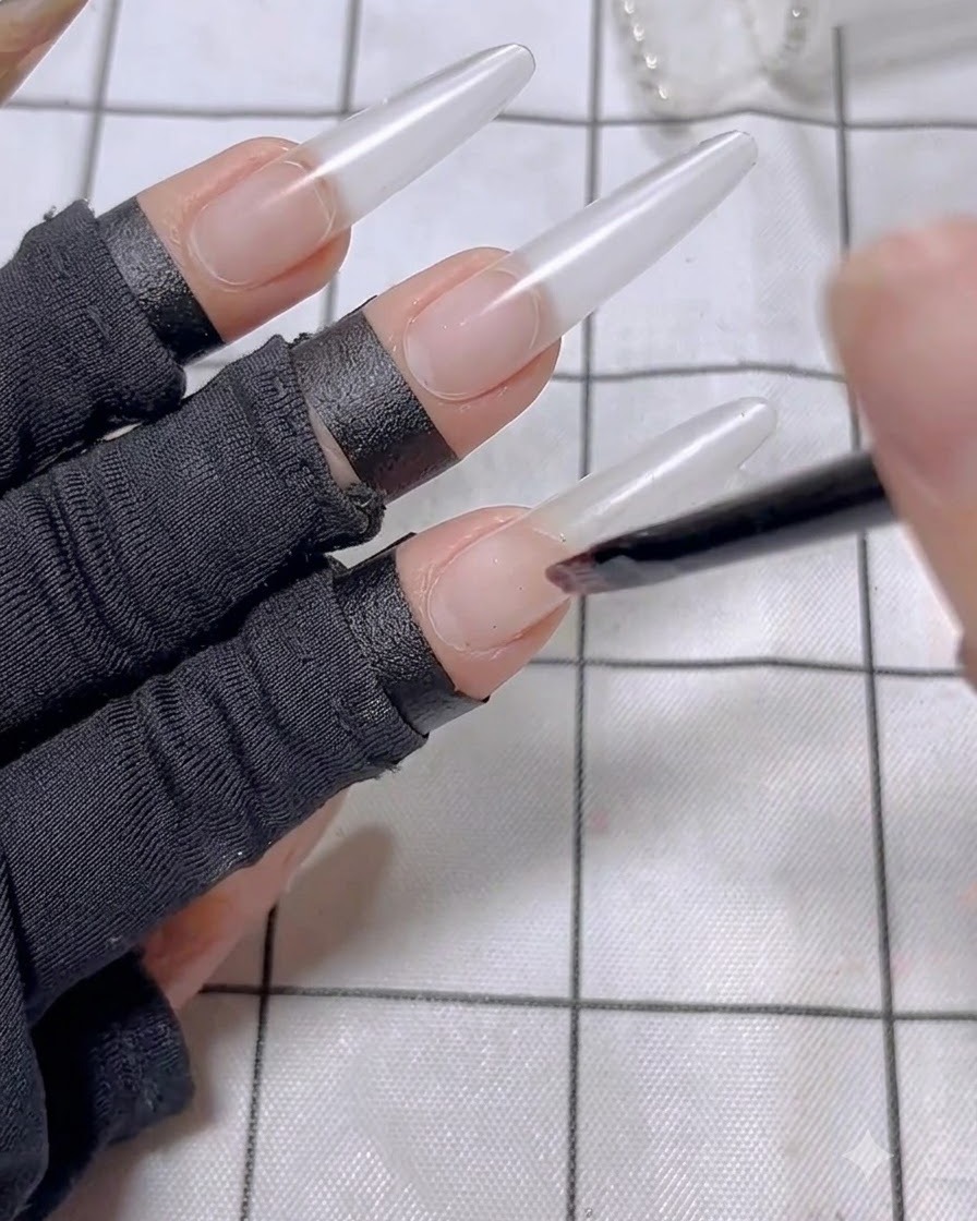 A nail brush applying a thin layer of nude base polish to the index finger nail extension.