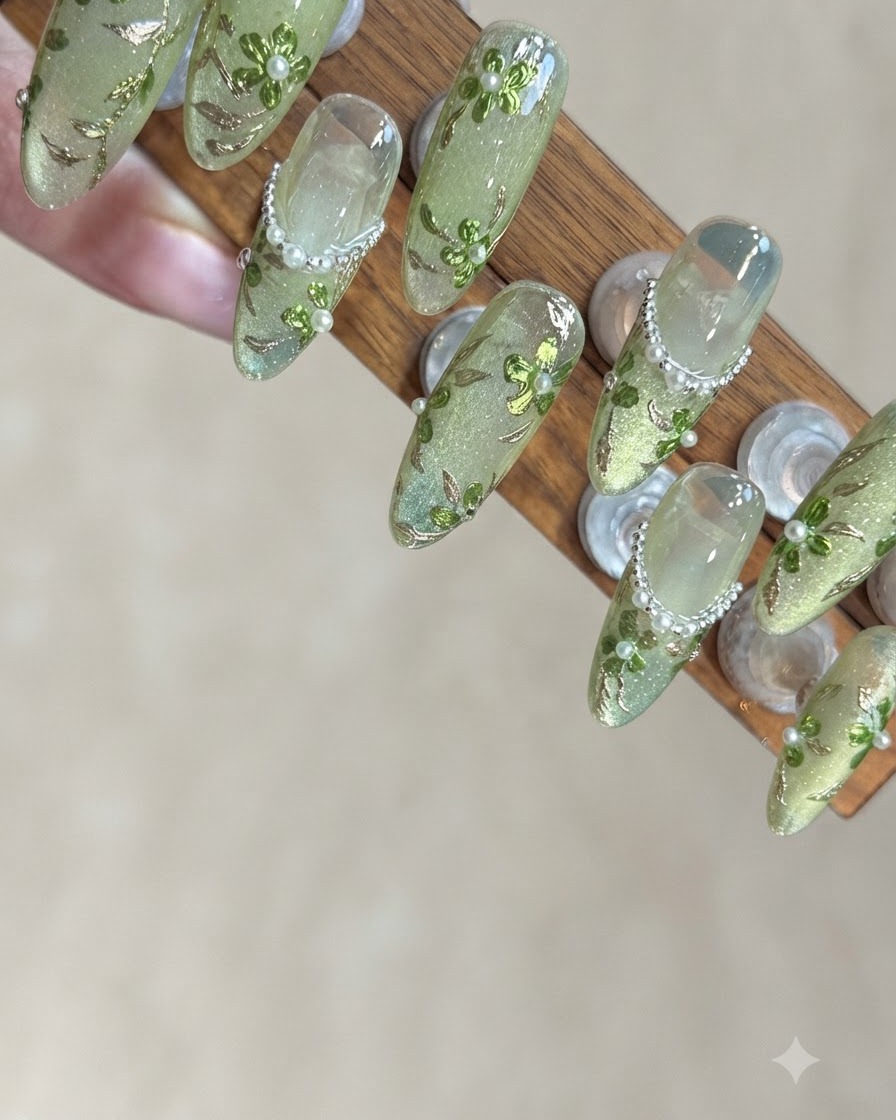 A close-up view of a manicurist applying tiny silver beads along the lower curve of a green glitter nail tip.