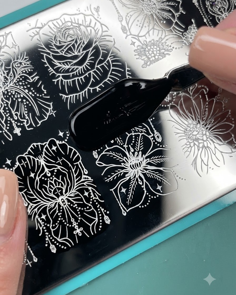 A brush applicator applying black stamping polish across an engraved floral pattern on a metal stamping plate.