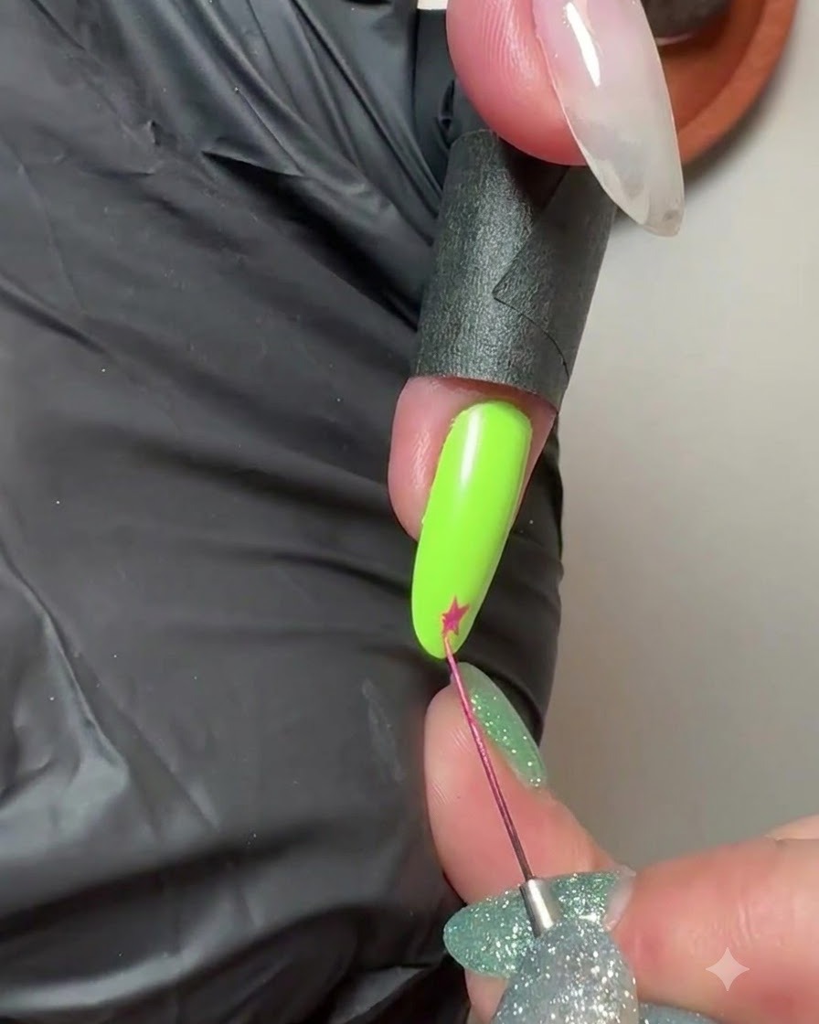 A fine liner brush drawing a small pink star on the tip of a neon green manicured nail.