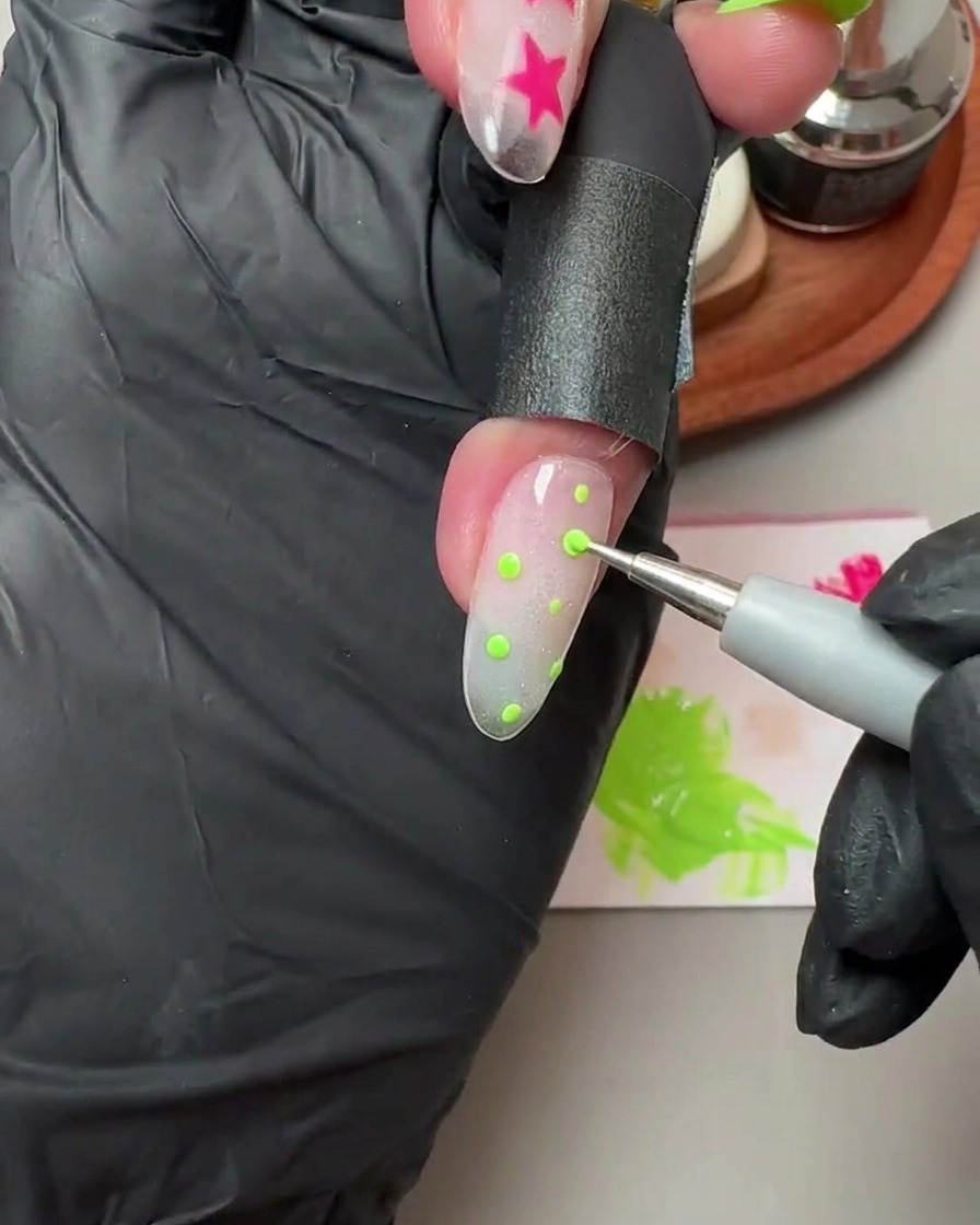 A dotting tool placing small green polka dots onto a translucent glitter and cat-eye gel nail.